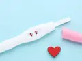 Biały test ciążowy z dwoma czerwonymi paskami, różowa nakładka i czerwone serduszko na niebieskim tle.