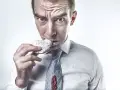 Mężczyzna w białej koszuli i krawacie je pączka, ubrudzony cukrem pudrem i dżemem.