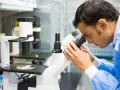 Mężczyzna w niebieskim fartuchu laboratoryjnym patrzy przez mikroskop.