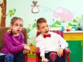 Dwoje dzieci w fotelikach, dziewczynka w okularach i chłopiec w muszce, na tle kolorowej ściany z rysunkami.
