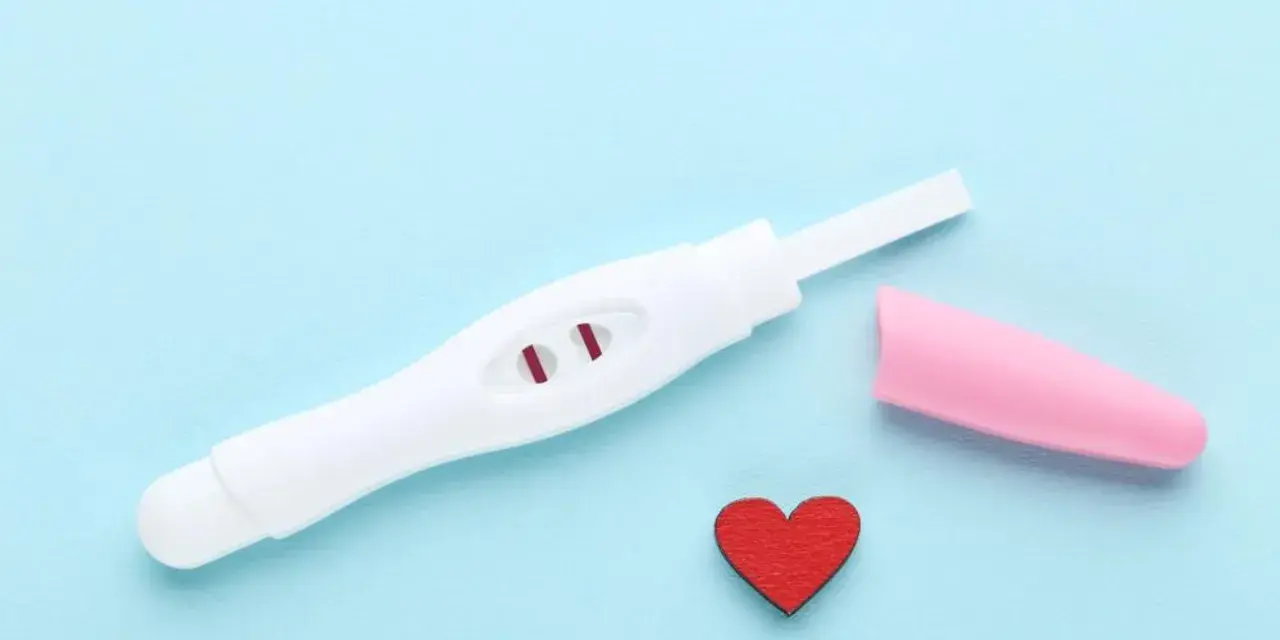 Biały test ciążowy z dwoma czerwonymi paskami, różowa nakładka i czerwone serduszko na niebieskim tle.