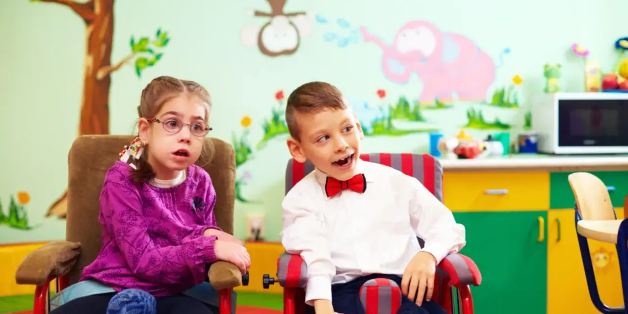 Dwoje dzieci w fotelikach, dziewczynka w okularach i chłopiec w muszce, na tle kolorowej ściany z rysunkami.