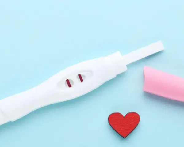 Biały test ciążowy z dwoma czerwonymi paskami, różowa nakładka i czerwone serduszko na niebieskim tle.