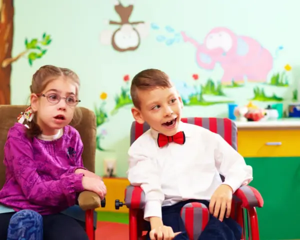 Dwoje dzieci w fotelikach, dziewczynka w okularach i chłopiec w muszce, na tle kolorowej ściany z rysunkami.