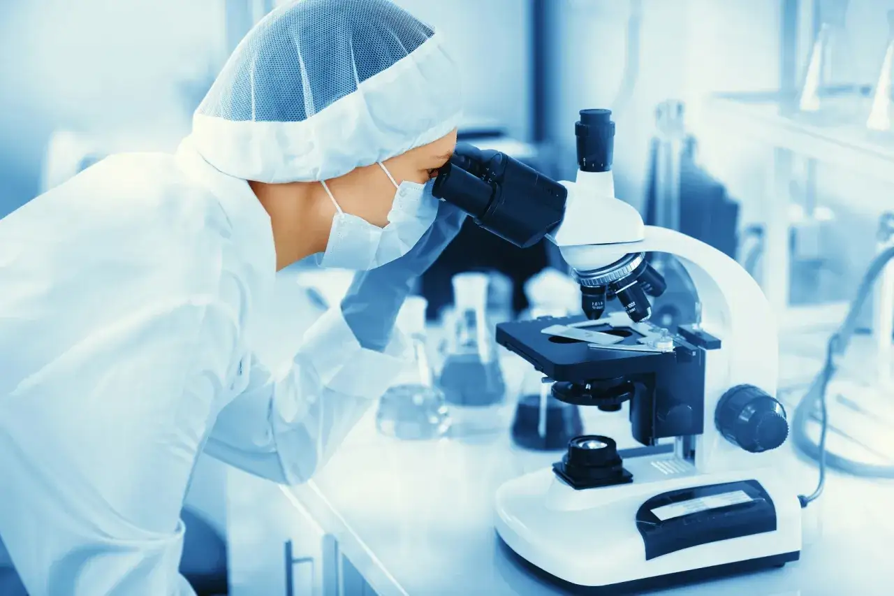 Naukowczyni w masce i czepku wykonuje badanie histopatologiczne pod mikroskopem w laboratorium.