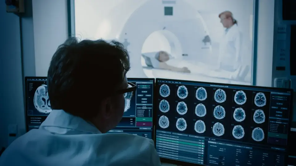 Lekarz analizuje obrazy mózgu na monitorze, gdzie widać wyraźny kontrast między tkankami. W tle pacjent leży w aparacie MRI.